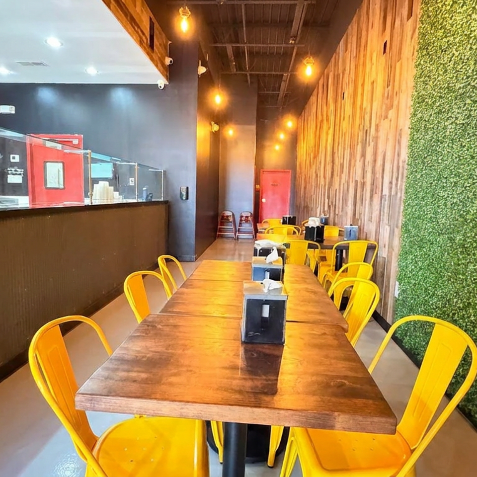 Bright casual restaurant dining area with wooden tables, yellow metal chairs, greenery wall, and open kitchen counter.